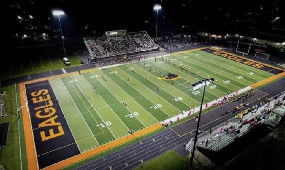 A night football game on the Little Eagles field, with "EAGLES" painted in the end zones, players on the turf and spectators behind the sideline.